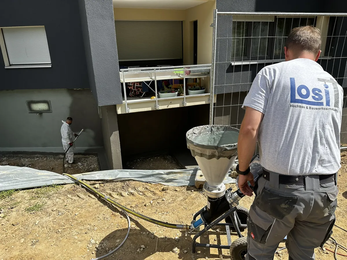Zwei Mitarbeiter von Losi Bau auf einer Baustelle: einer bedient ein Mörtelgerät, der andere bearbeitet den Gebäudesockel im offenen Aushub